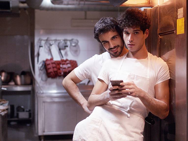 Zwei männlich gelesene Personen stehen in einer Restaurantküche an einen Schrank gelehnt. Die vordere Person hält ein Handy in der Hand, während die hintere ihr über die Schulter schaut und in die Kamera lächelnd.